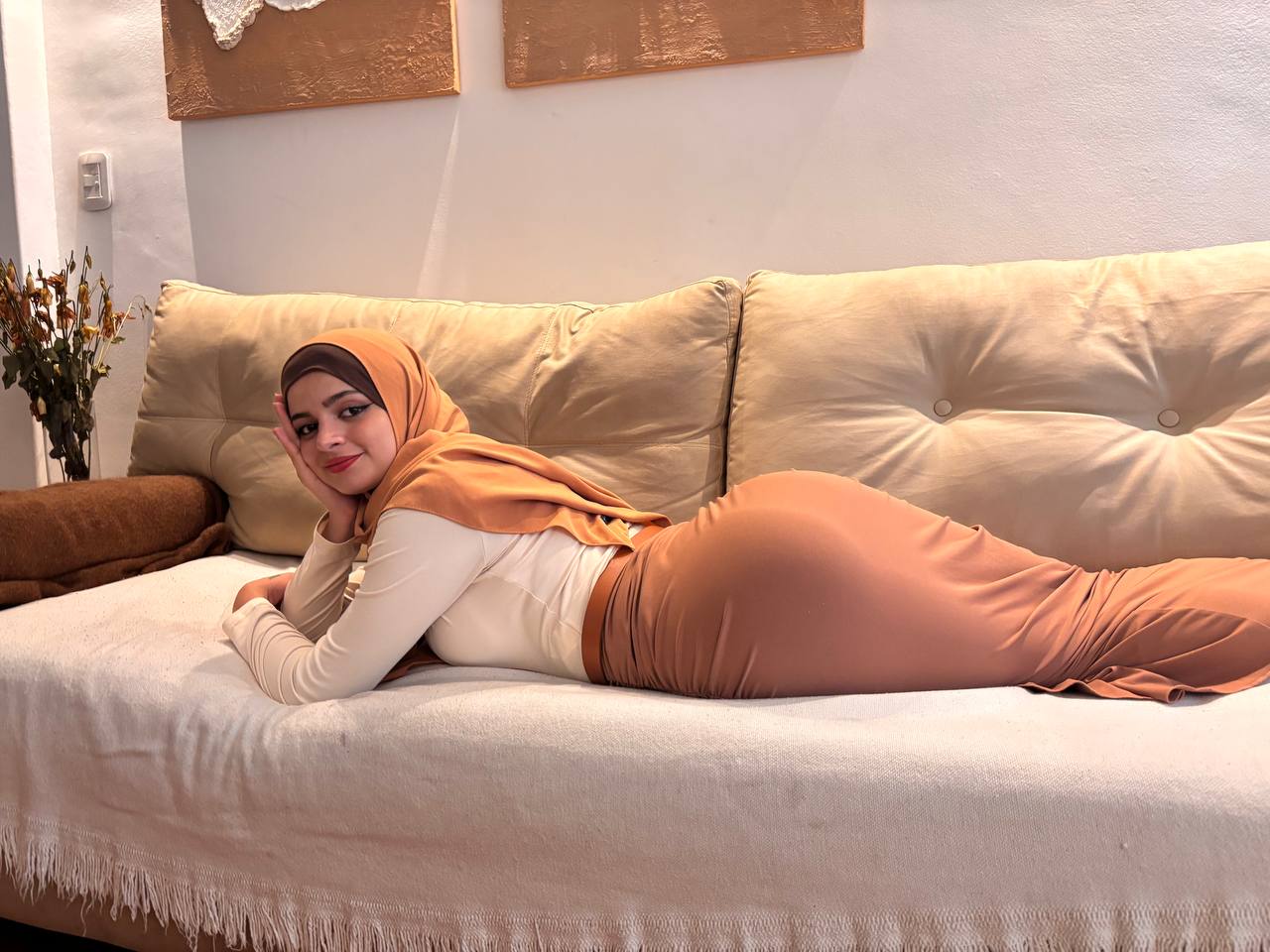 Woman in a peach hijab and light outfit lying on her side on a cream sofa, propped on one elbow and smiling.
