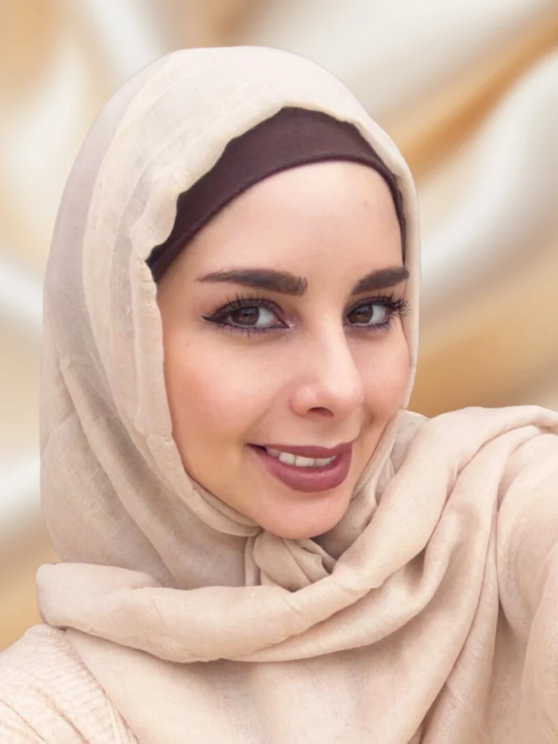 Close-up of a smiling woman wearing a beige hijab and brown headband, looking at the camera against a soft, warm background.