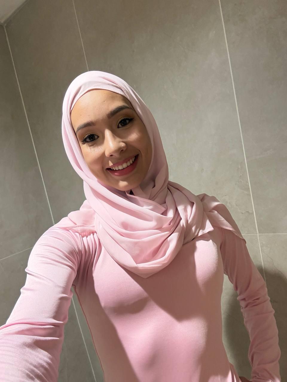 Young woman wearing a pale pink hijab and matching long-sleeve top, smiling at the camera in a tiled bathroom backdrop.