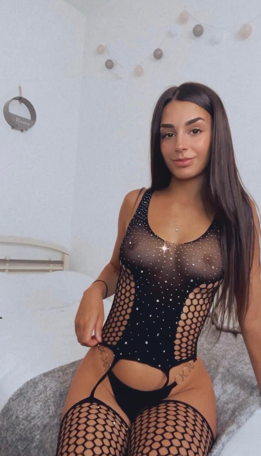 Young woman posing on a bed in a sparkly black mesh bodysuit with fishnet sides.