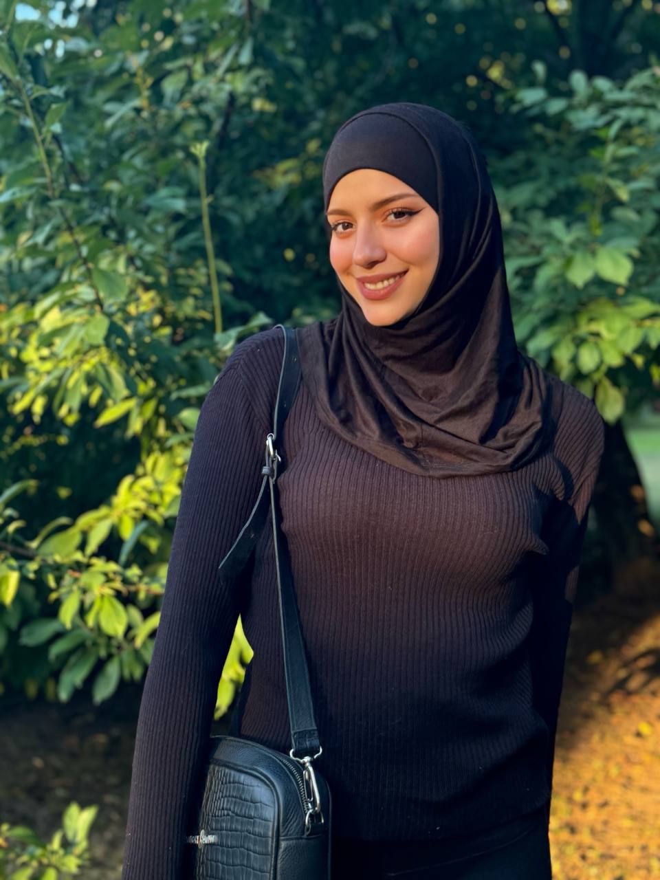 Smiling woman wearing a black hijab and sweater, standing in a leafy outdoor setting with a black crossbody bag.