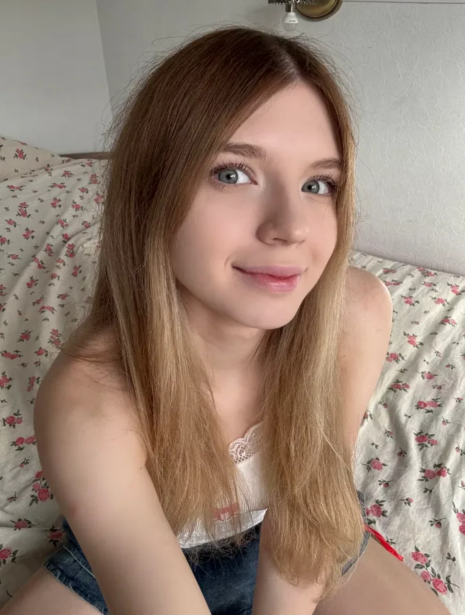 Young woman with long light brown hair and blue eyes smiling at the camera on a floral bedspread outdoors indoors.