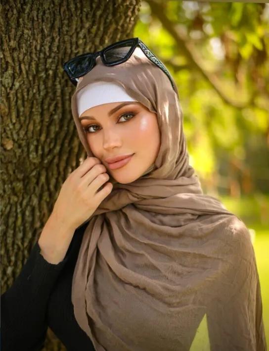 Woman wearing a beige hijab and white inner headband, with sunglasses resting on top of her head, posing beside a tree in a sunny park.
