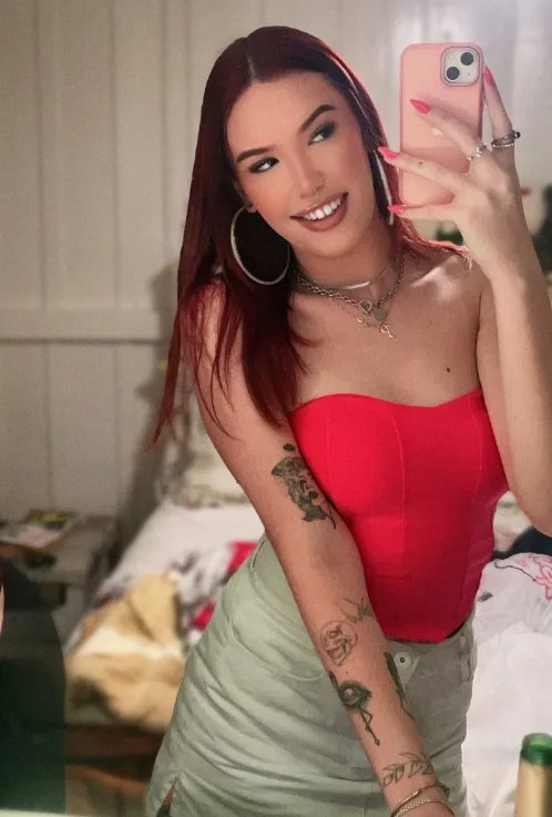 Woman in a mirror selfie wearing a red strapless top and light green skirt, tattoos on her left arm, smiling at the camera.
