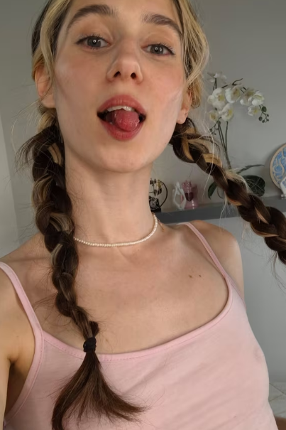 Young woman with two braids wearing a pink tank top and pearl necklace, sticking out her tongue playfully; orchid in the background.