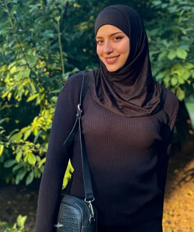 Smiling woman wearing a black hijab and ribbed sweater, standing outdoors with green plants behind her, carrying a black crossbody bag.