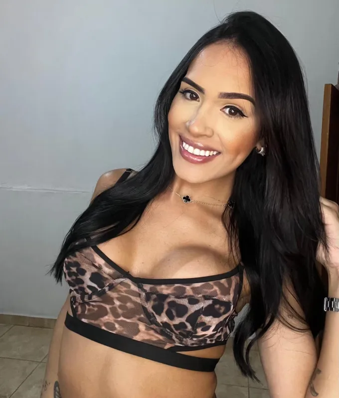 Smiling woman with long dark hair wearing a leopard-print bralette and a delicate necklace, posing for a selfie indoors.