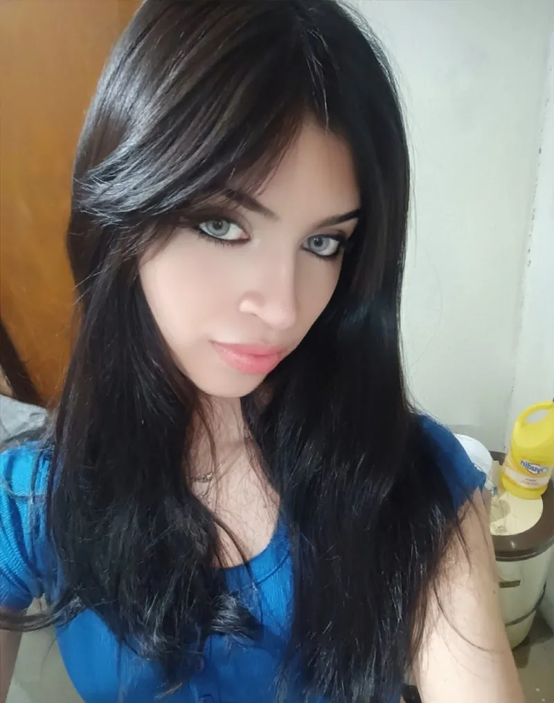 Close-up of a young woman with long dark hair and blue eyes, wearing a blue top, looking at the camera.