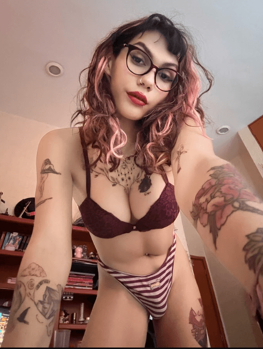 Selfie of a person with pink hair and glasses wearing a burgundy bra and striped panties, tattoos on arms and chest, in a bedroom setting.